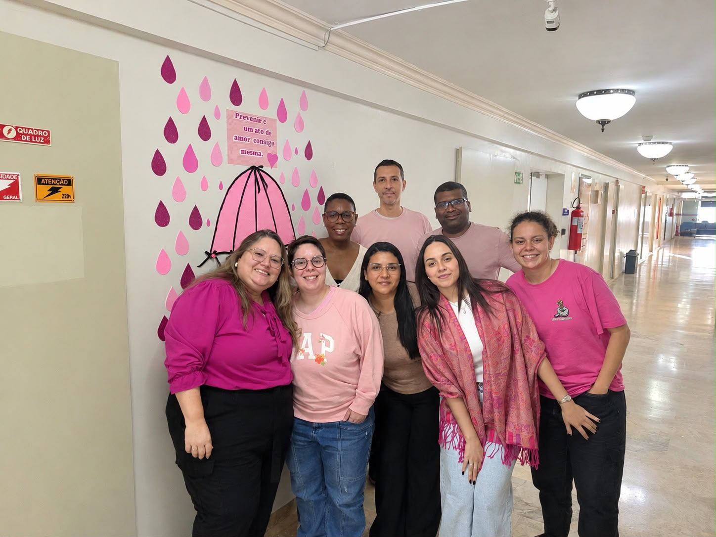 Grupo de oito pessoas posa sorrindo em um corredor, com roupas em tons variados e rosa, em alusão ao Outubro Rosa. Ao lado, um painel decorativo traz um guarda-chuva rosa e a frase: Prevenir é um ato de amor consigo mesma.