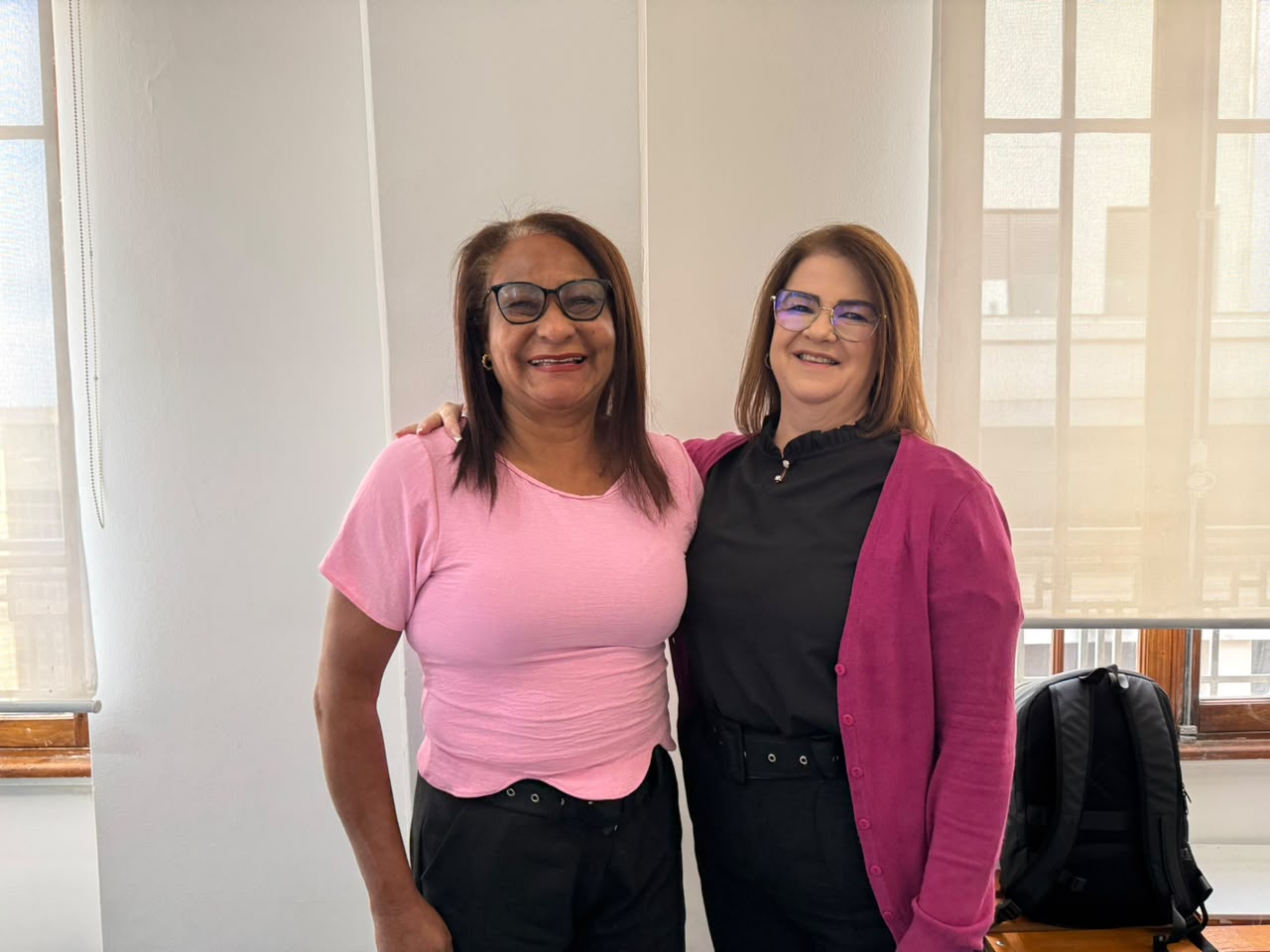 Duas mulheres posam abraçadas pelos ombros e sorrindo em um escritório. Uma veste camiseta rosa e a outra usa casaco de tricô rosa-escuro sobre blusa preta.