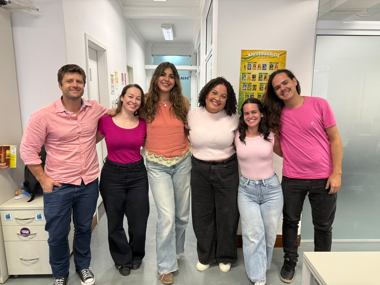 Grupo de seis pessoas, quatro mulheres e dois homens, posa sorrindo de braços dados em um corredor. Todos vestem roupas em tons de rosa e salmão, em apoio ao Outubro Rosa. Ao fundo, há um cartaz de aniversários de 2026.