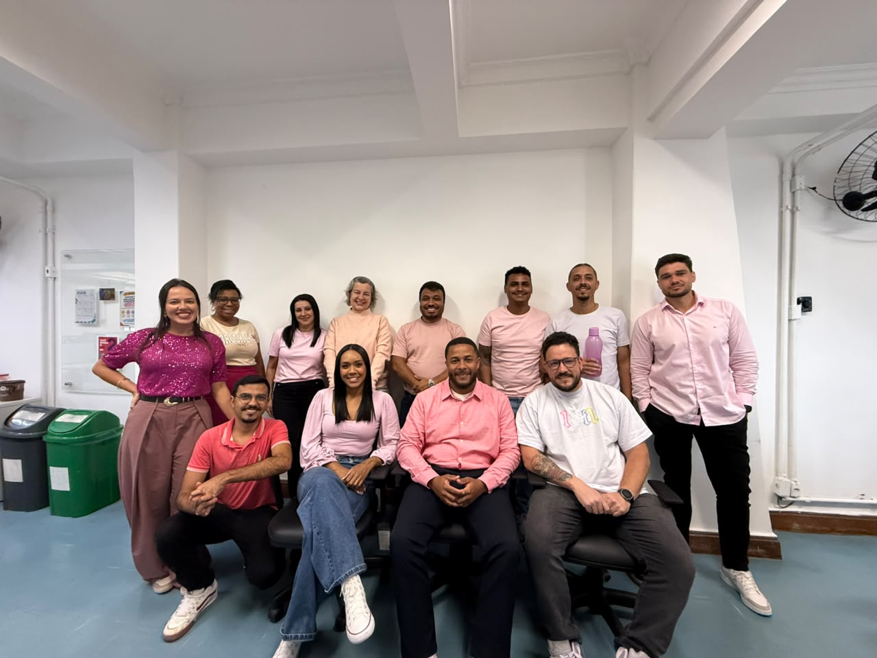 Grupo de doze pessoas posa sorrindo em um escritório. Quatro pessoas estão sentadas ou agachadas à frente e oito estão em pé atrás. O grupo veste roupas em tons de rosa, magenta, salmão e branco.