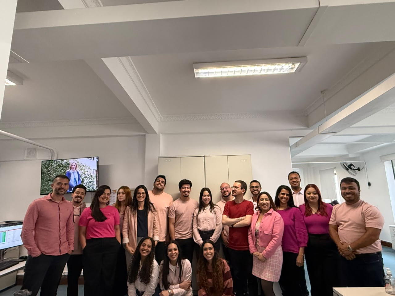 Grupo de dezesseis pessoas posa sorrindo em um ambiente corporativo. Três mulheres estão agachadas à frente e as demais pessoas estão em pé atrás. A maioria veste roupas em tons de rosa e vermelho.