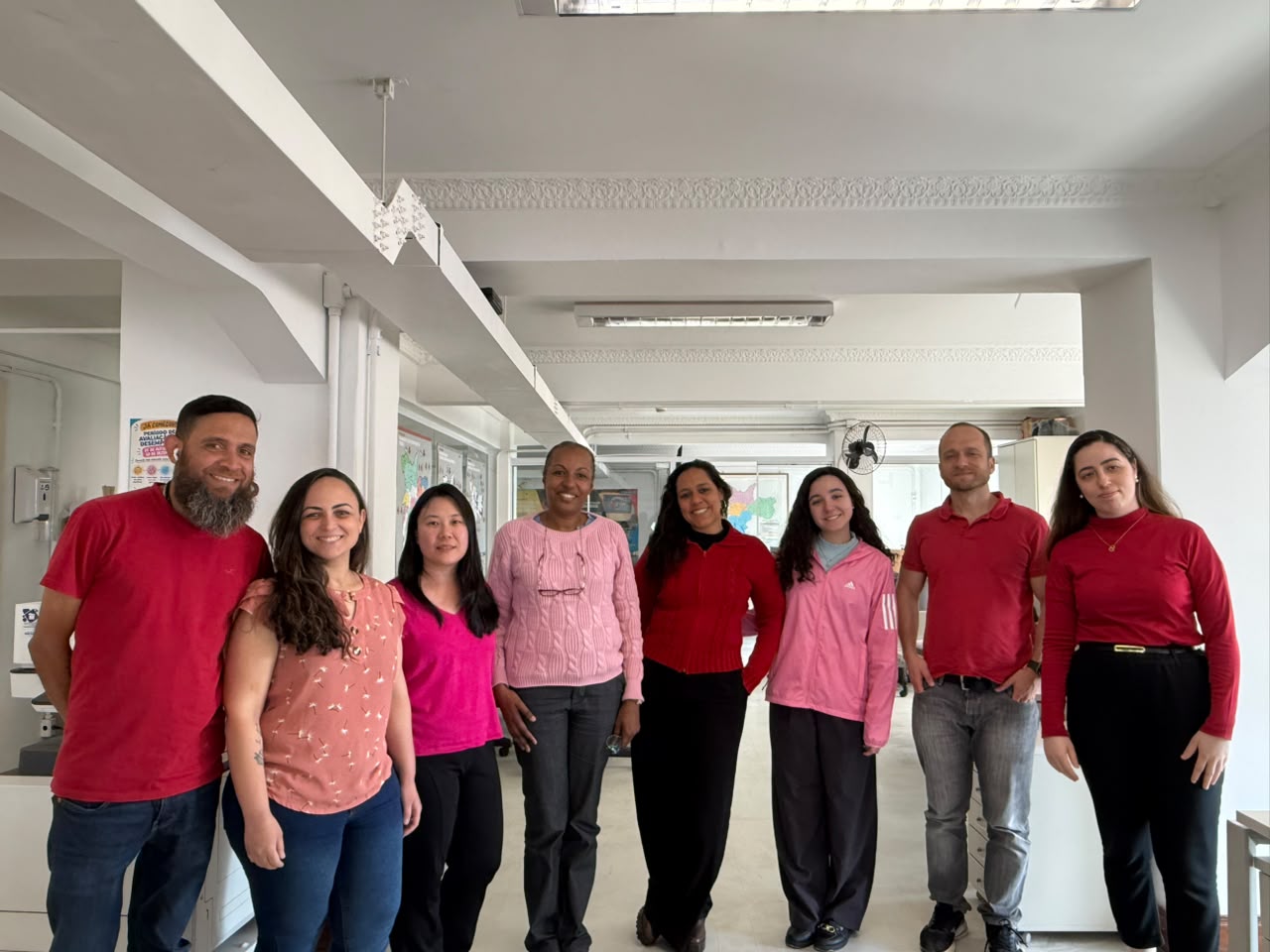 Oito pessoas, cinco mulheres e três homens, posam lado a lado em um escritório. Todas vestem blusas ou camisetas em tons de rosa e vermelho, em apoio à campanha do Outubro Rosa.