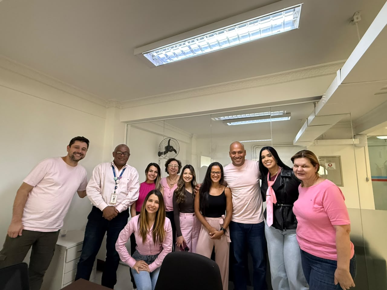Grupo de dez pessoas posa sorrindo em um escritório. A maioria veste roupas ou acessórios em tons de rosa, em alusão à campanha do Outubro Rosa.