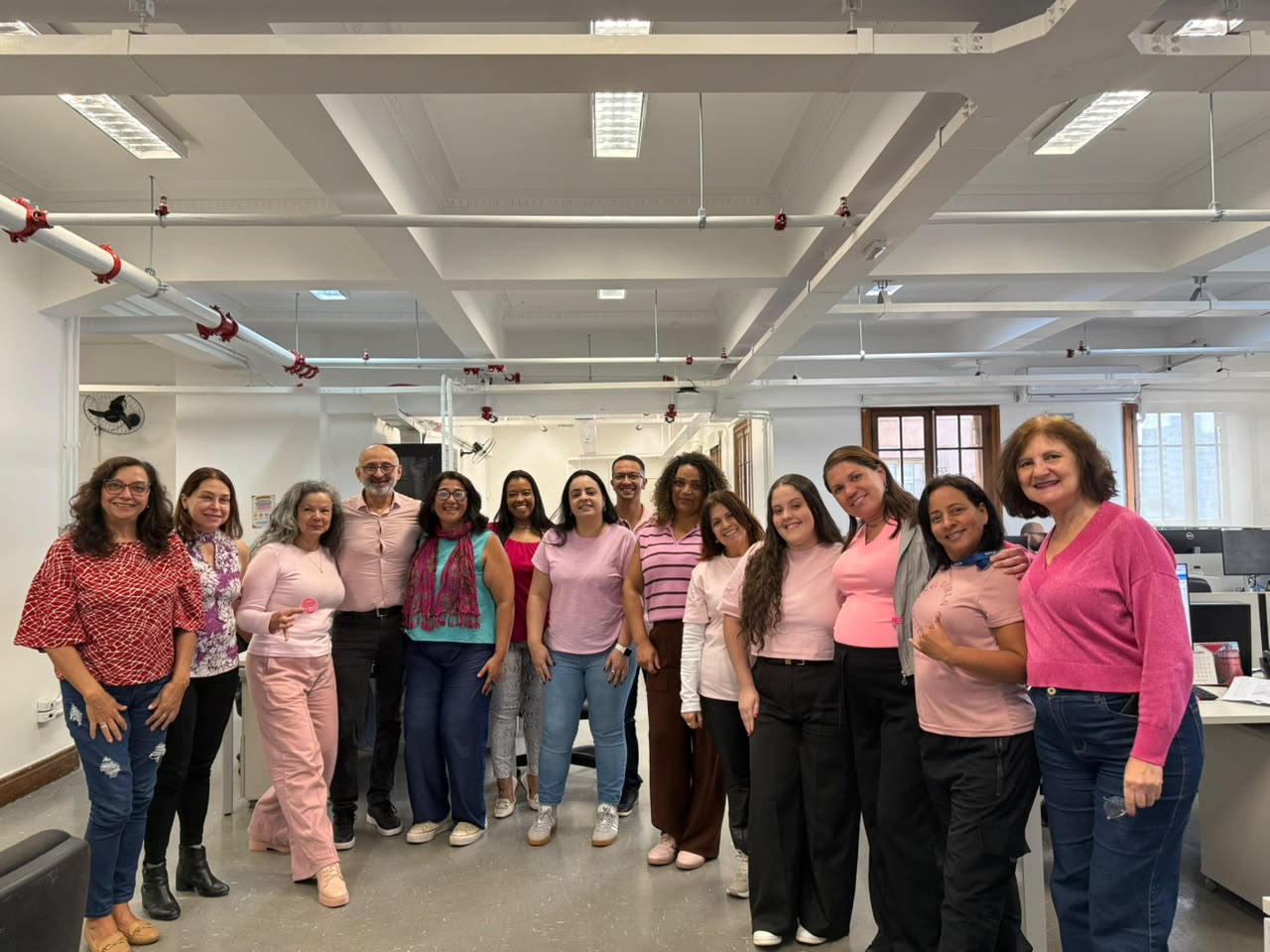 Grupo de catorze pessoas, entre mulheres e homens, posa em um amplo escritório. As pessoas vestem roupas em tons de rosa e vermelho, em apoio ao Outubro Rosa.