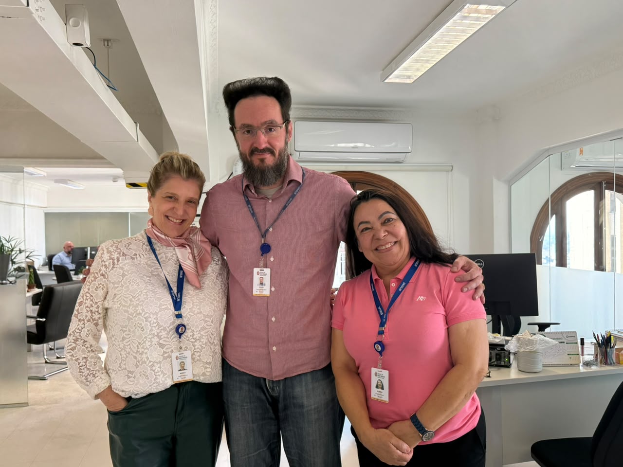 Duas mulheres e um homem posam sorrindo em um escritório. As três pessoas usam crachás corporativos e peças de roupa em tons de rosa, em alusão ao Outubro Rosa.