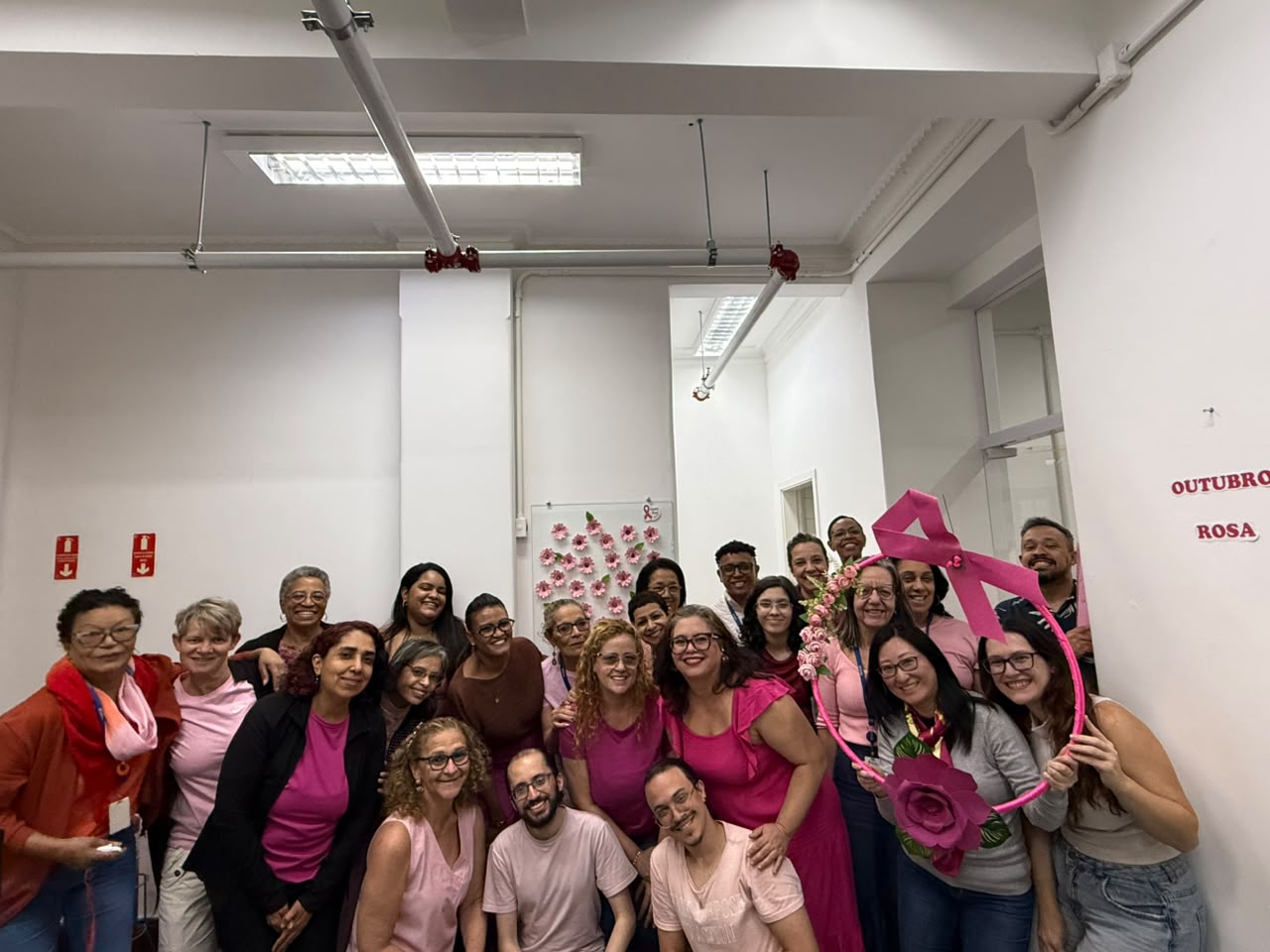 Grupo de vinte e duas pessoas posa sorrindo em um escritório. Duas mulheres seguram um arco decorativo rosa com laço e flores. Ao fundo, há um painel com flores de papel e a inscrição Outubro Rosa.