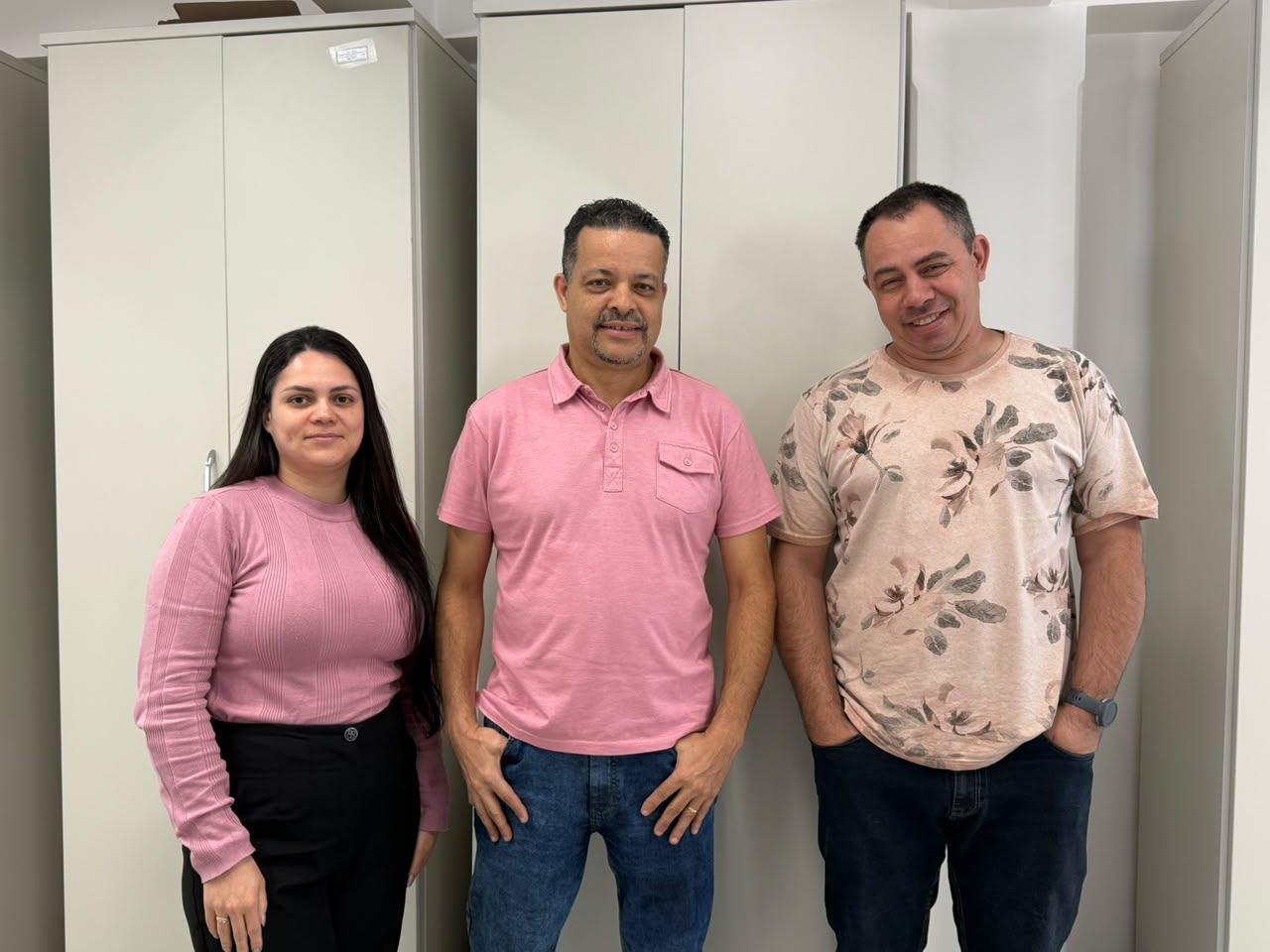 Uma mulher e dois homens posam sorrindo lado a lado em um ambiente corporativo. Os três vestem roupas em tons de rosa, em alusão à campanha do Outubro Rosa. Ao fundo, há armários bege.