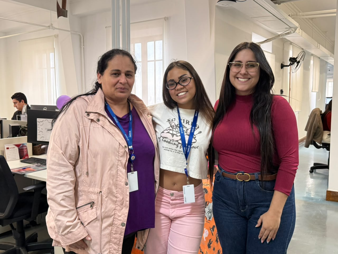 Três mulheres posam sorrindo em um escritório. Elas vestem jaqueta rosa-claro, camiseta branca com estampa e blusa em tom vinho. Ao fundo, há estações de trabalho e um ventilador de parede.