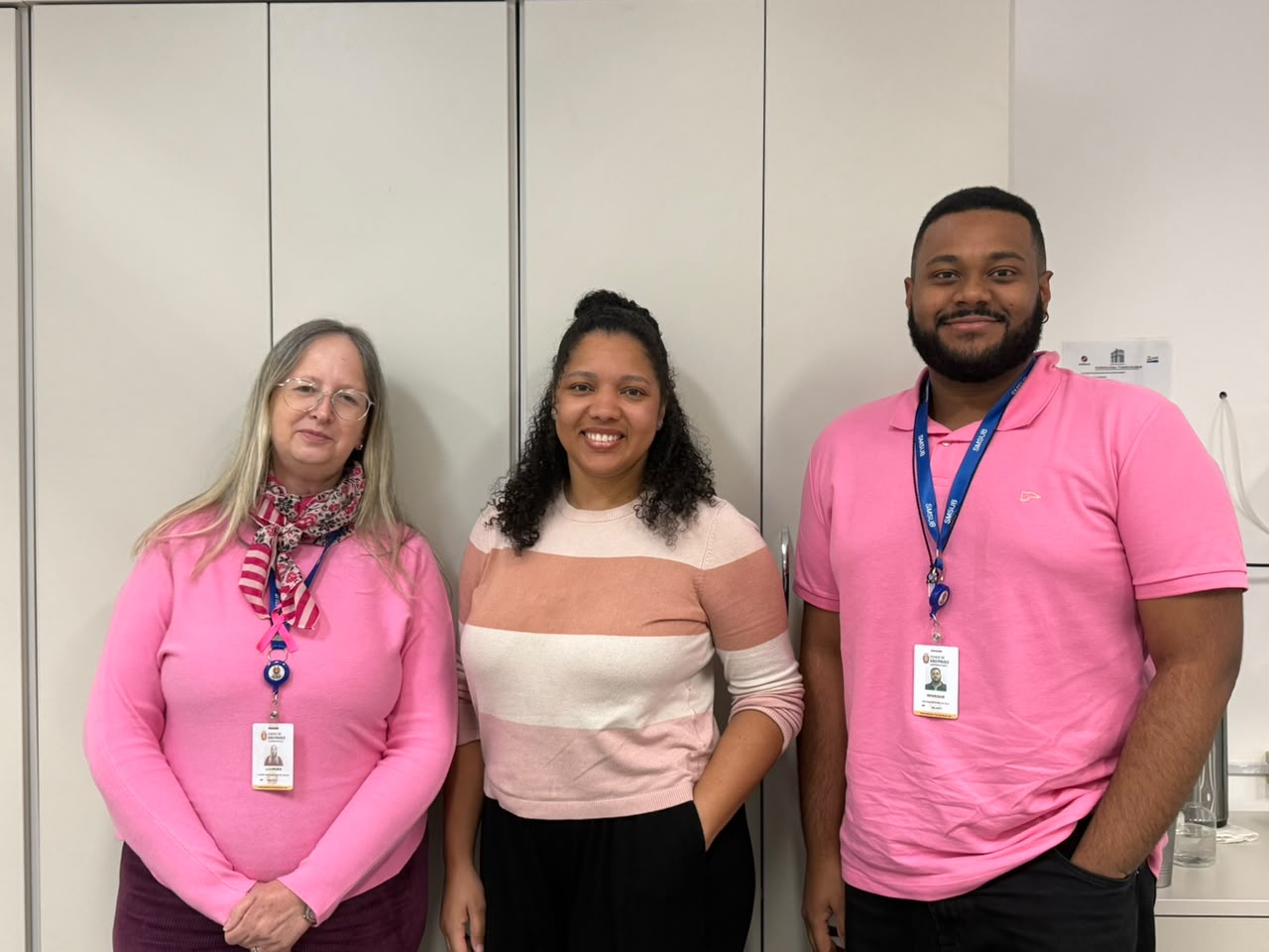 Duas mulheres e um homem posam sorrindo em um ambiente corporativo, usando roupas em tons de rosa. Uma das mulheres usa um laço rosa da campanha no peito.