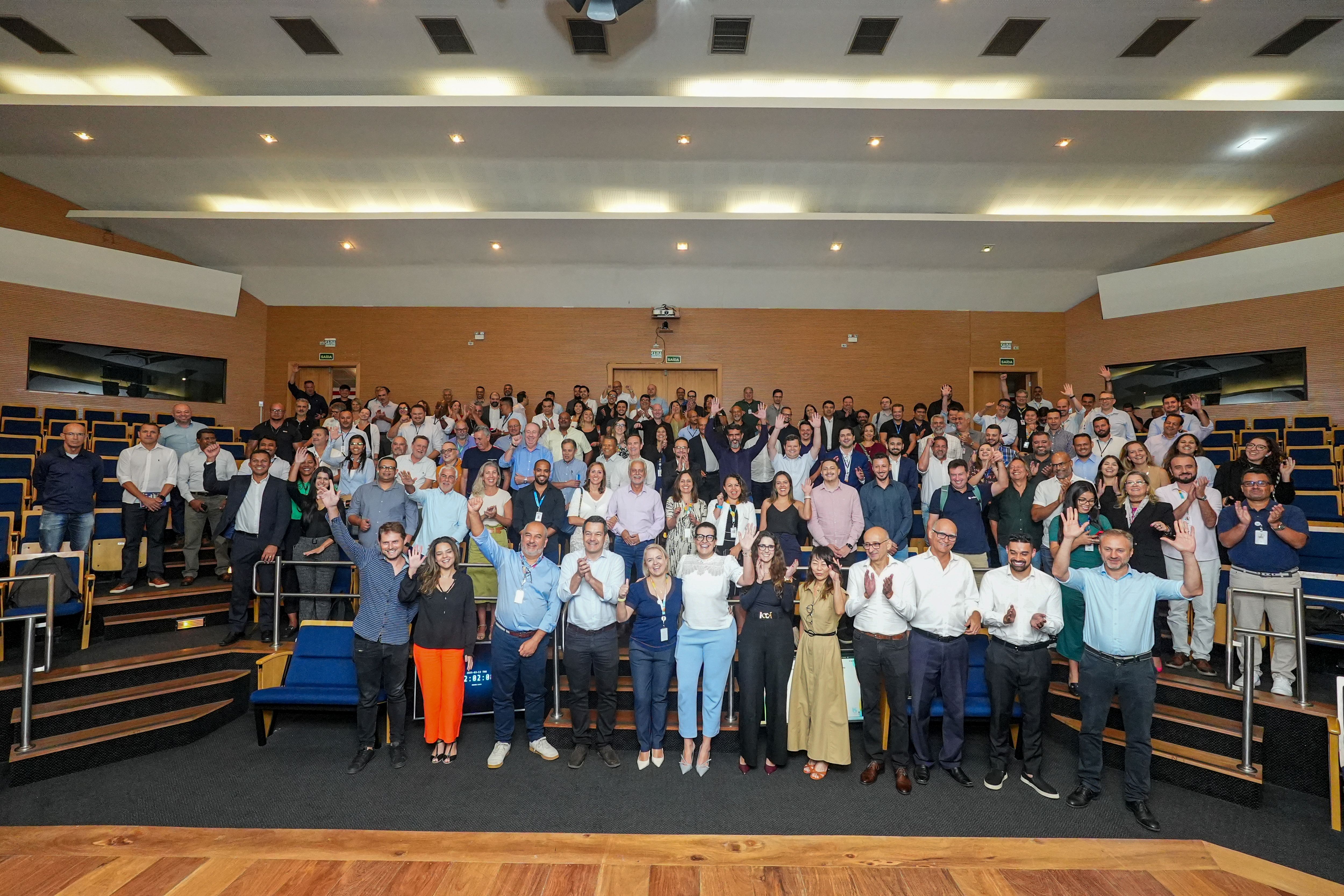 A imagem mostra um grande grupo de pessoas reunidas em um auditório moderno, posando para uma foto coletiva. Os participantes estão distribuídos em diferentes níveis das cadeiras estofadas e muitos deles sorriem, levantam os braços ou fazem gestos de comemoração. O ambiente é bem-iluminado, com um design contemporâneo, paredes em tons neutros e um teto com iluminação embutida. A diversidade de expressões e posturas sugere um momento de celebração ou encerramento de um evento, conferência ou reunião corporativa.