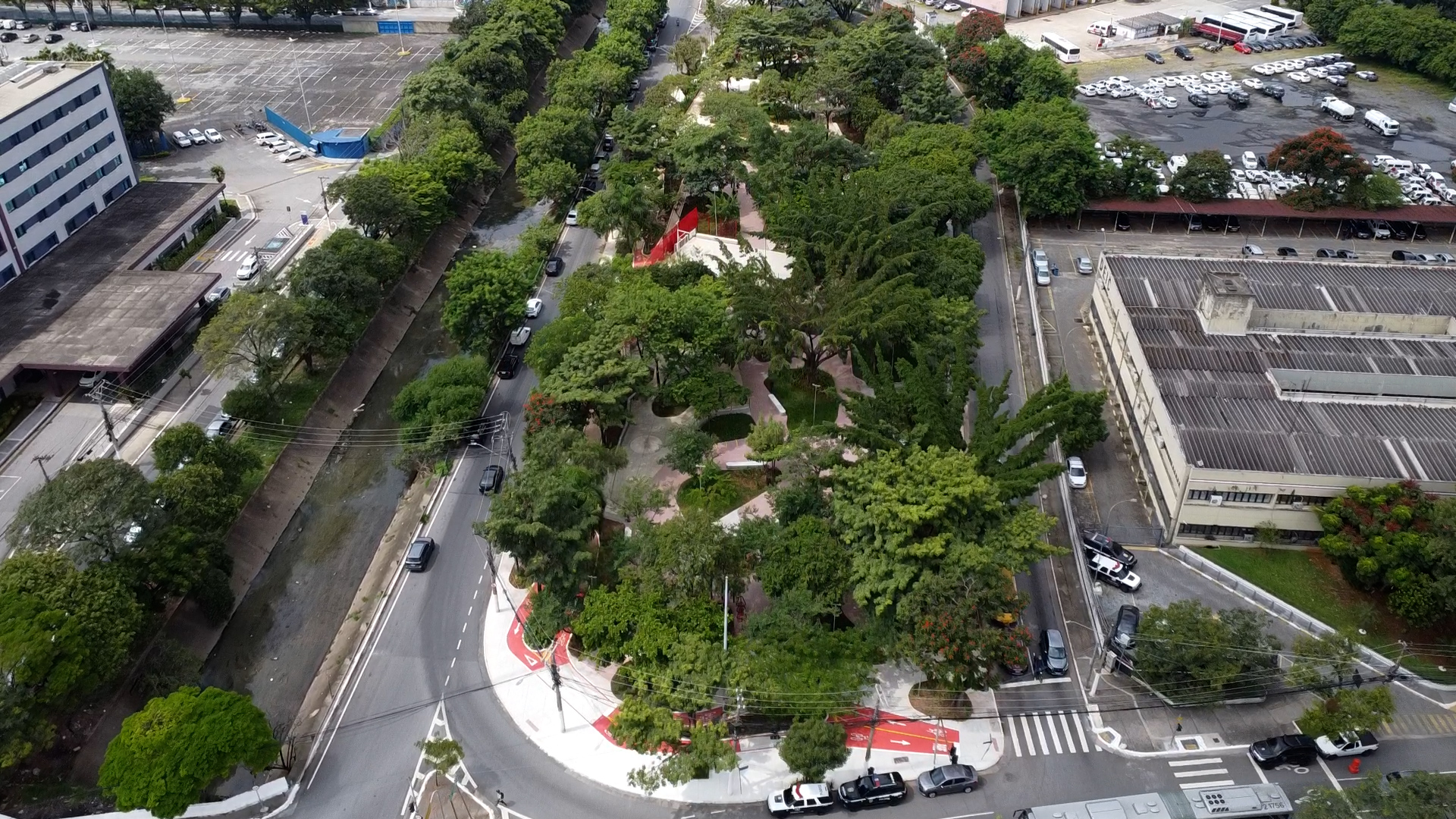 Imagem aérea de uma praça urbana repleta de árvores e vegetação, cercada por ruas movimentadas com carros estacionados e em circulação. O espaço verde conta com caminhos pavimentados, bancos e uma estrutura vermelha no centro, que pode ser um monumento ou um equipamento público. Há ciclovias demarcadas ao redor da praça, destacadas em vermelho. Nas proximidades, há prédios comerciais e institucionais, além de um grande estacionamento com diversos veículos. Um pequeno canal de drenagem passa ao lado da praça, acompanhado por uma via com tráfego moderado.