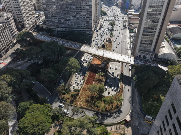 #PraCegoVer Uma imagem aérea em plano geral, tirada durante o dia, mostra uma paisagem urbana densa, em uma cidade como São Paulo, onde destaca-se um viaduto para pedestres que atravessa uma via de tráfego intenso. O viaduto, em tom de branco, tem formato arredondado, é curvilíneo e é sustentado por colunas, que não são vistas na imagem. A passarela do viaduto é branca com bordas amarelas e está repleta de pessoas caminhando, cruzando de um lado para o outro. No meio da imagem, duas vias de tráfego opostas, com vários veículos transitando, passam por baixo do viaduto. Do lado esquerdo da imagem, há um aglomerado de edifícios de prédios de diversos tamanhos, no lado direito da imagem, há prédios mais modernos e altos. Na parte superior da imagem, mais vias de tráfego e prédios. Na parte inferior, há árvores grandes que cobrem a via de pedestres e as laterais do viaduto, na parte central de baixo, mais vias de trânsito, e na extremidade direita inferior, a lateral de um edifício moderno.