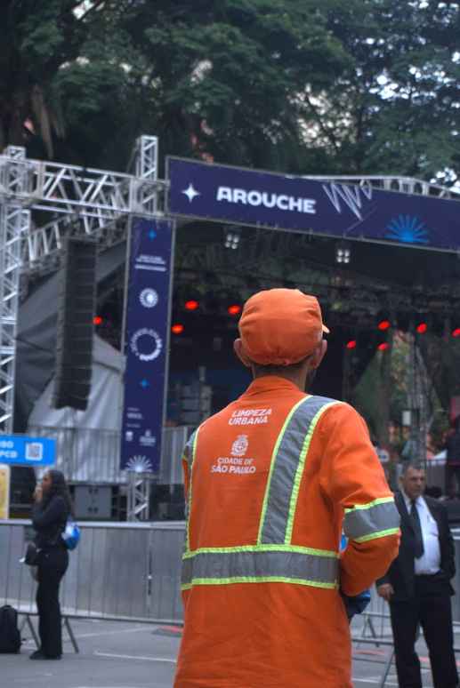 Um homem de boné e uniforme laranja, com faixas reflexivas cinzas e a inscrição "LIMPEZA URBANA CIDADE DE SÃO PAULO" nas costas, está de costas para o observador. Ele parece estar em um evento ao ar livre, possivelmente um festival ou show, dado o cenário com uma estrutura de palco metálica, caixas de som e uma faixa azul com o nome "AROUCHE" e elementos gráficos. As luzes vermelhas do palco são visíveis ao fundo. Outras pessoas, incluindo uma mulher de pé à esquerda e um homem de terno à direita, podem ser vistas em segundo plano. O ambiente geral sugere uma área de evento urbano.