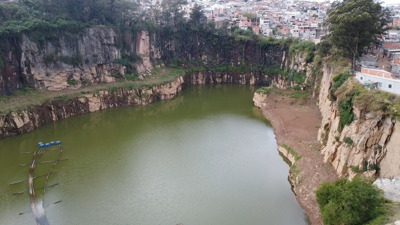 Foto aérea de uma pedreira desativada, com um lago de água verde no centro. As paredes da pedreira são rochosas e irregulares, com tons de cinza e bege. No canto inferior esquerdo, há um poste de madeira com fios azuis pendurados. No canto superior direito, casas e prédios urbanos são visíveis, indicando a proximidade da cidade. A vegetação é esparsa, com algumas árvores e arbustos crescendo nas bordas da pedreira. O céu está nublado, com tons de cinza e branco.