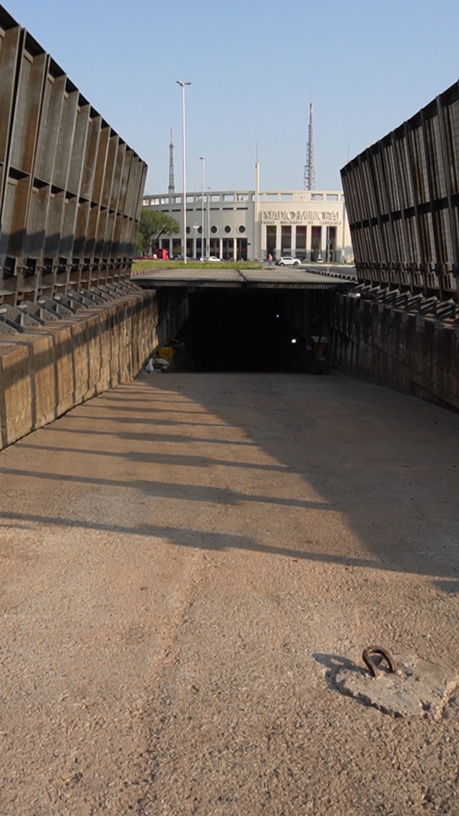 Foto de um túnel de concreto com duas estruturas metálicas elevadas em ambos os lados, criando uma passagem sombreada. As estruturas metálicas têm painéis verticais com espaços vazados, formando um padrão geométrico. O chão do túnel é de concreto claro, com uma sombra escura no centro, sugerindo a entrada para um espaço subterrâneo. Ao fundo, um prédio branco com colunas e janelas retangulares é visível, sob um céu azul claro. A luz do sol ilumina a cena, destacando as texturas do concreto e das estruturas metálicas.