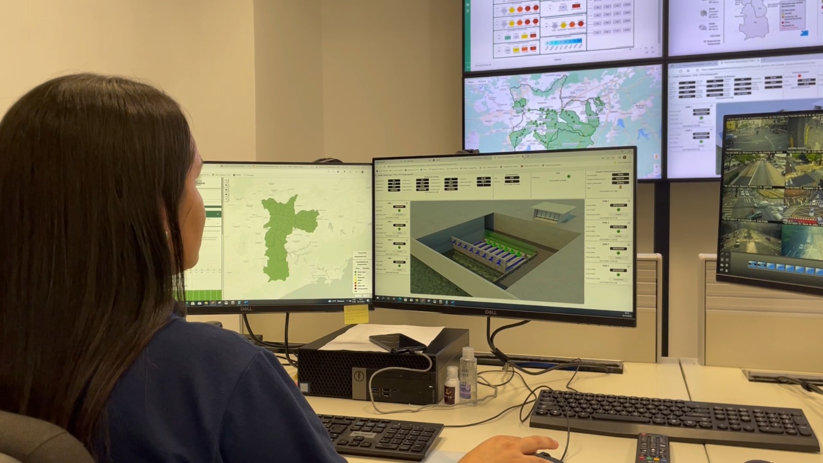 Foto de uma mulher de costas, sentada em frente a dois monitores de computador em uma sala de controle. Ela tem cabelos longos e escuros e veste uma camisa azul. Os monitores exibem mapas, gráficos e imagens de câmeras de segurança. Ao fundo, há outros monitores maiores na parede, mostrando informações semelhantes. A sala é clara e organizada, com equipamentos eletrônicos e teclados visíveis. A mulher parece estar trabalhando ou monitorando as telas.