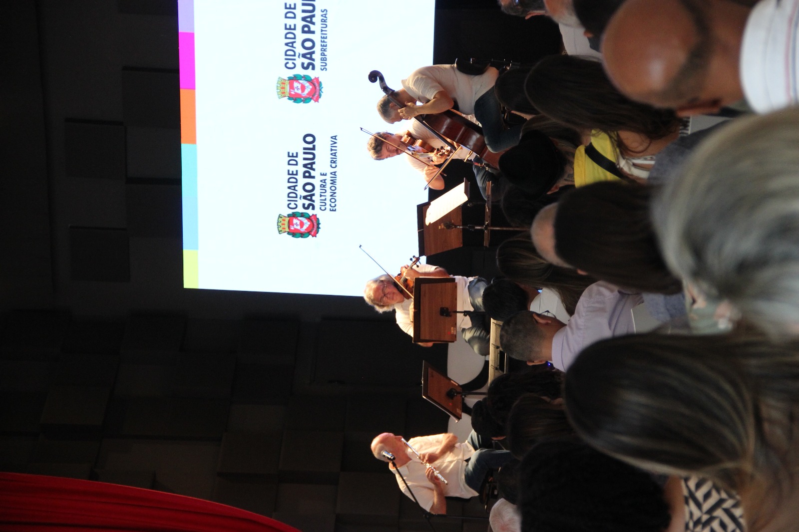  Foto de um palco com quatro músicos tocando instrumentos de corda e sopro para uma plateia. Ao fundo, uma tela de LED com os logos da "CIDADE DE SÃO PAULO". A plateia está sentada de costas para a câmera, assistindo à apresentação. A iluminação é suave e o ambiente sugere um evento cultural.
